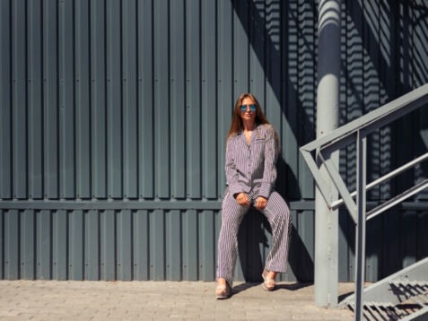 Junge Frau mit Sonnenbrille in blau-weiß gestreiftem Pyjama lehnt an eine Wand aus grauem festem Wellblech.