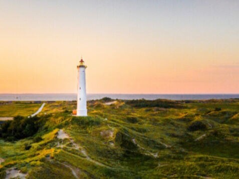 Weißer Leuchtturm auf grünen Dünen bei Sonnenuntergang mit Dorf und Meer im Hintergrund.