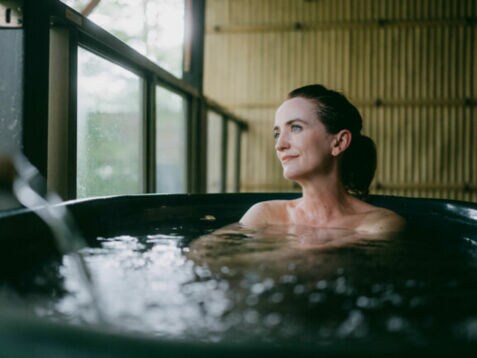 Eine Frau sitzt in einem Thermalbad aus dunklem Stein in einem japanischen Spa mit Bambuswand und schaut zufrieden durch ein Fenster hinaus.