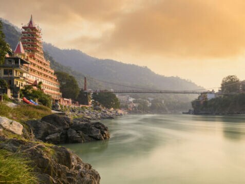 Fluss mit hängender Fußgängerbrücke und mehrstöckigem Tempelgebäude am Ufer bei Sonnenuntergang in bergiger Landschaft