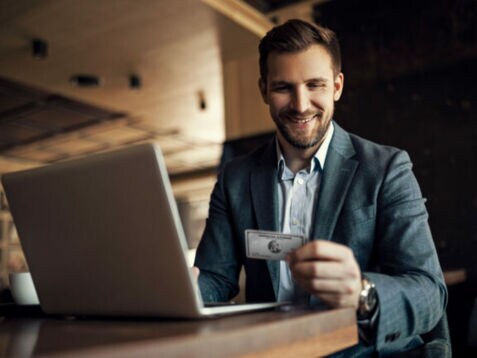 Ein lächelnder Geschäftsmann sitzt an einem Tisch vor einem Laptop und blickt auf eine silberne Platinum Kreditkarte von American Express in seiner Hand.