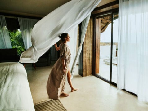 Eine Frau im langen Kleid und einem Weinglas in der Hand steht in einer Suite und blickt hinaus durch eine geöffnete Terassentür mit wehenden Gardinen.