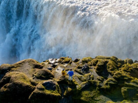 Zwei Personen stehen auf moosbedecktem Felsen vor einem großen Wasserfall mit starker Gischtentwicklung.