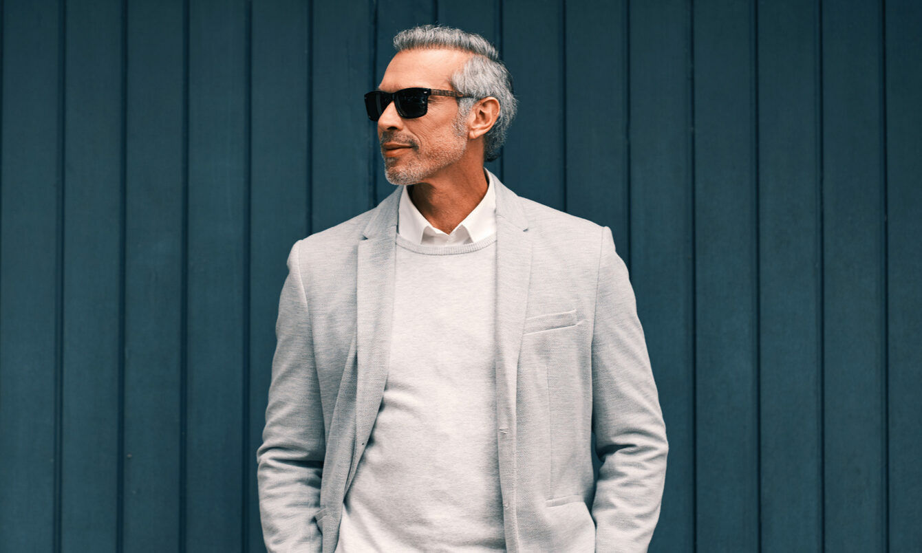 Mann vor einer blauen Holzwand mit weißem Pullover und hellem Anzug mit Sonnenbrille