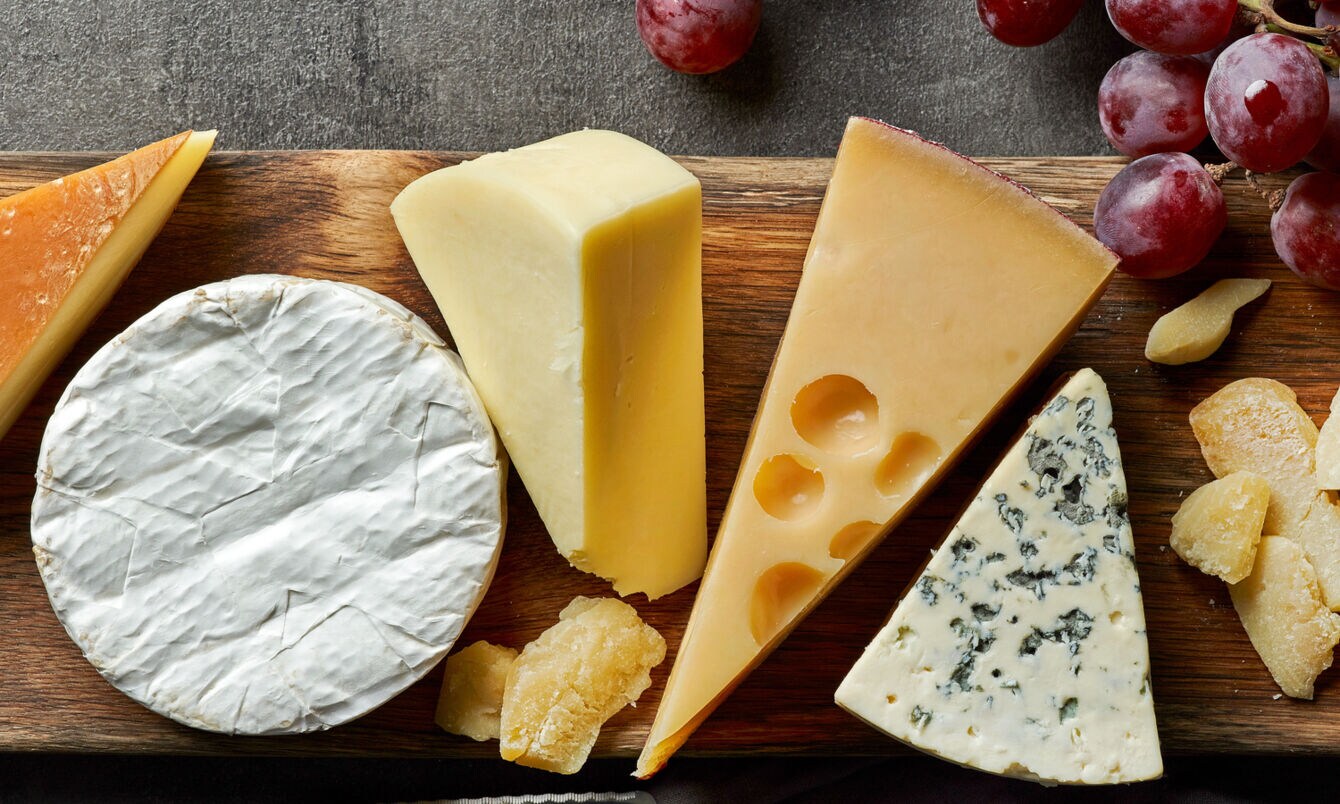 Verschiedene Käsesorten auf einem Holzbrett, daneben ein Messer und rote Trauben