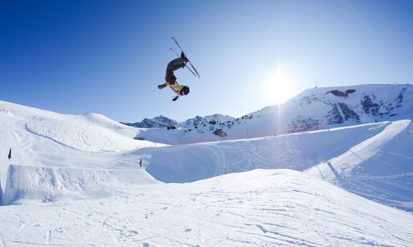 Eine Person, die mit Ski einen Salto in der Luft macht, im Hintergrund ein schneebedeckter Berg