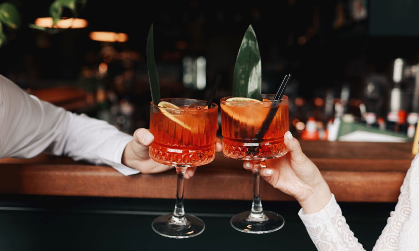 Zwei Spritz-Getränke mit Orangenscheiben in den Händen zweier Personen beim Anstoßen an einem Bartresen.