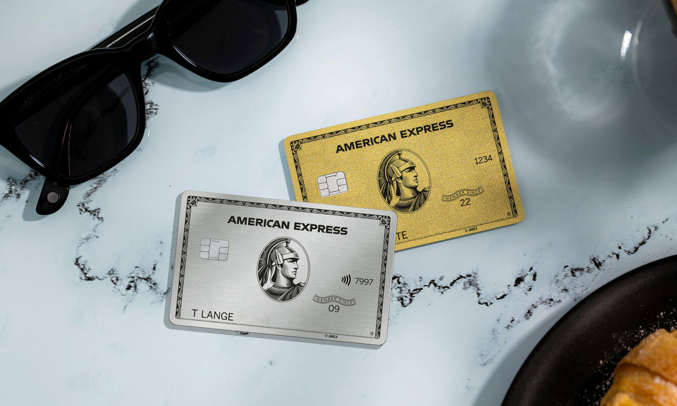 Eine silberne und eine goldene Kreditkarte von American Express liegen auf einem weißen Marmortisch neben einer schwarzen Sonnenbrille, einer Kaffeetasse und einem Teller mit Gebäck.