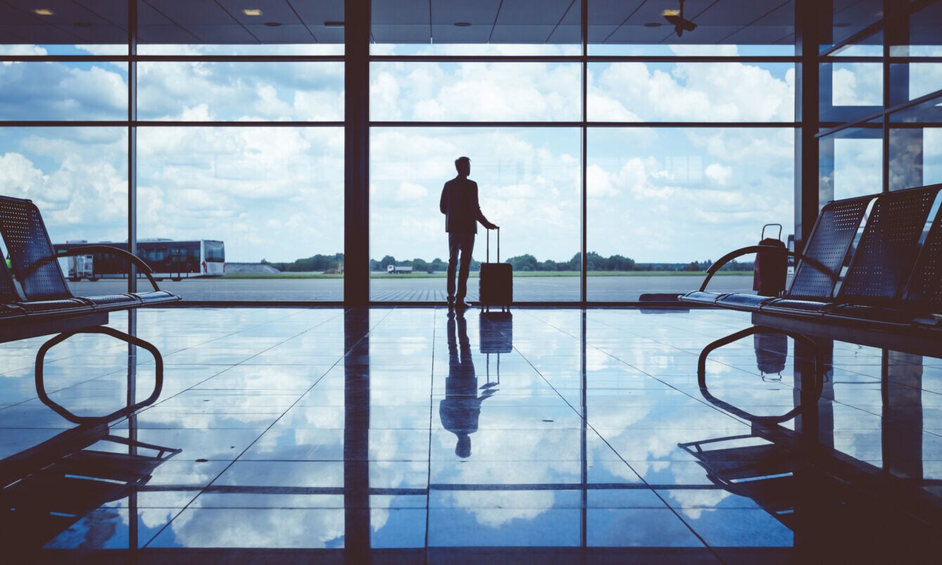 Rückenansicht einer Person, die in einer Flughafenhalle mit großer Glasfront am Fenster steht und einen Rollkoffer in einer Hand am Griff hält.