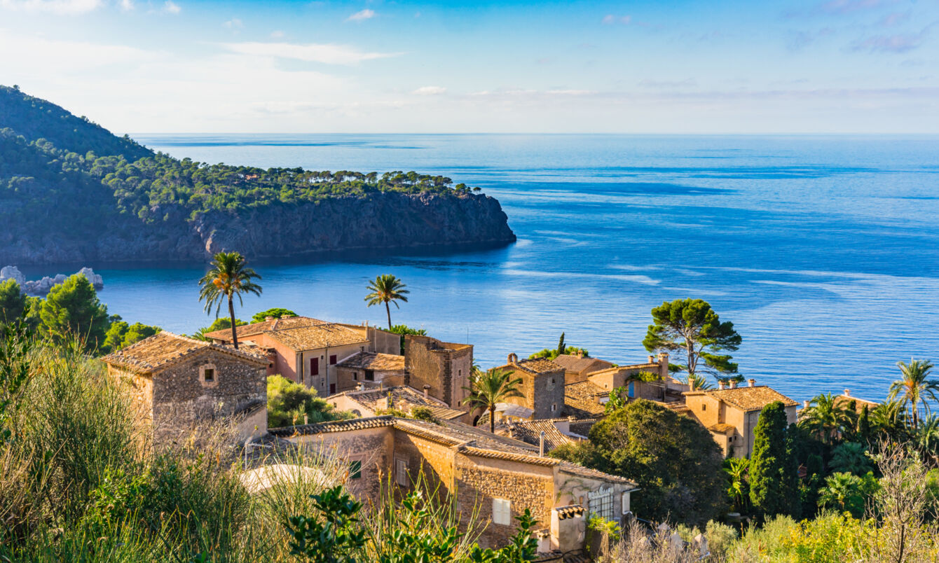 Ein altes Dorf an der Küste Mallorcas. Im Hintergrund eine grün überwachsene Landzunge, die in das Meer hineinragt.