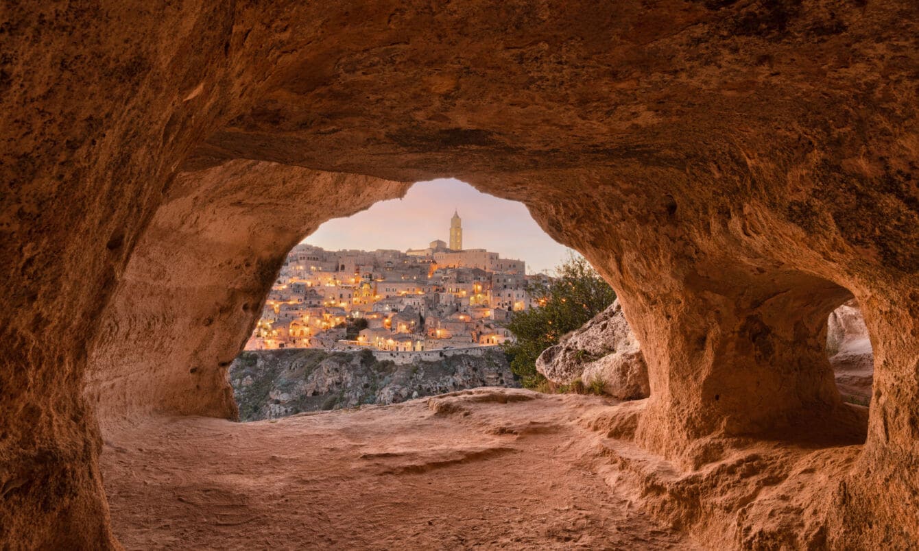 Blick auf die erleuchtete Stadt Matera aus einer Steingrotte im Vordergrund bei Dämmerung.