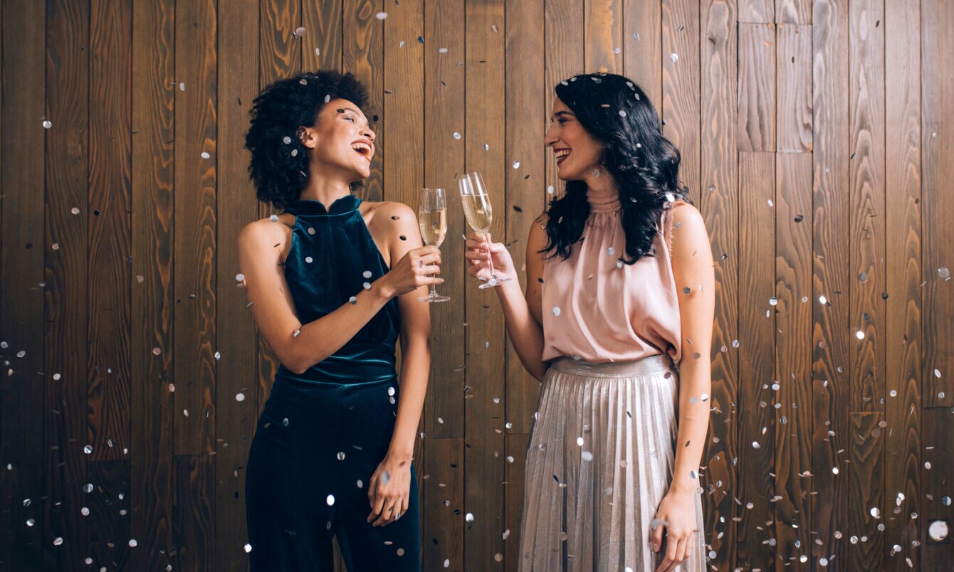 Zwei elegant gekleidete Frauen stehen im Konfettiregen vor einer Holzwand und stoßen mit Sektgläsern an.