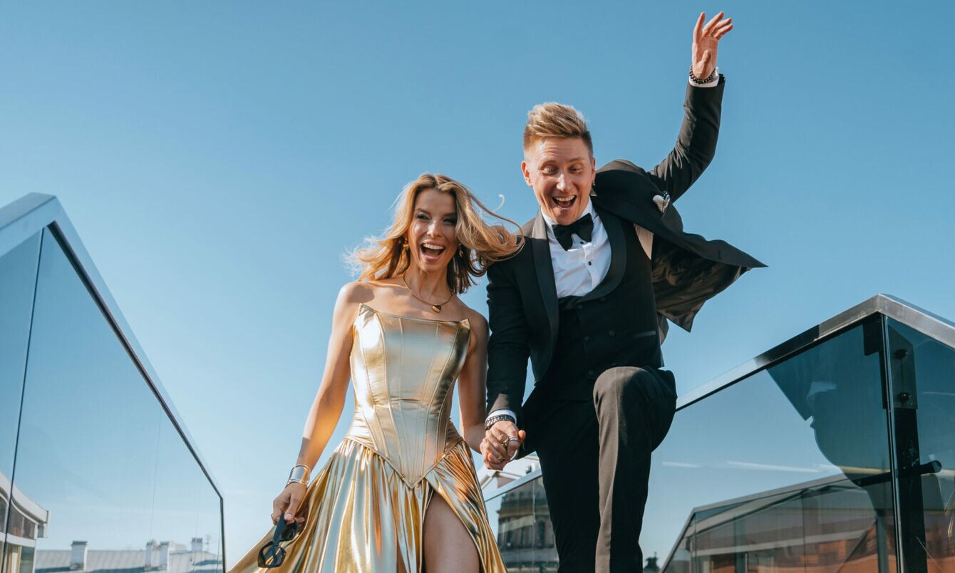 Ein lachendes Paar in Abendgarderobe läuft händchenhaltend in einem urbanen Umfeld.