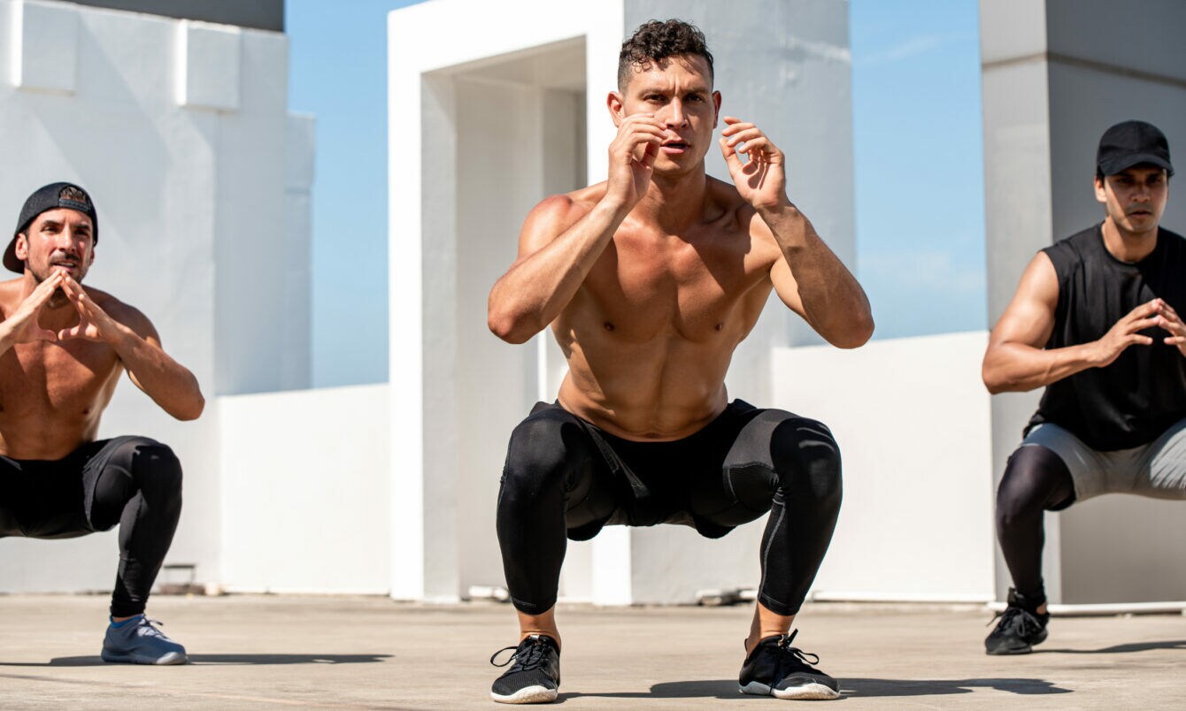 Drei Männer machen Kniebeugen im Freien vor weißer Architektur, zwei sind oben ohne, einer trägt ein schwarzes Shirt und eine Kappe