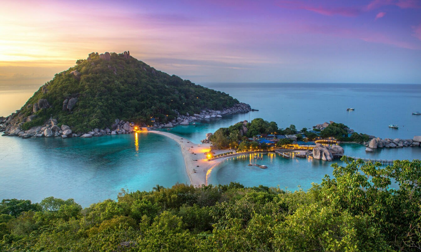 Blick auf eine tropische Insel mit bewaldetem Hügel, Sandbank und beleuchteten Gebäuden bei Sonnenuntergang