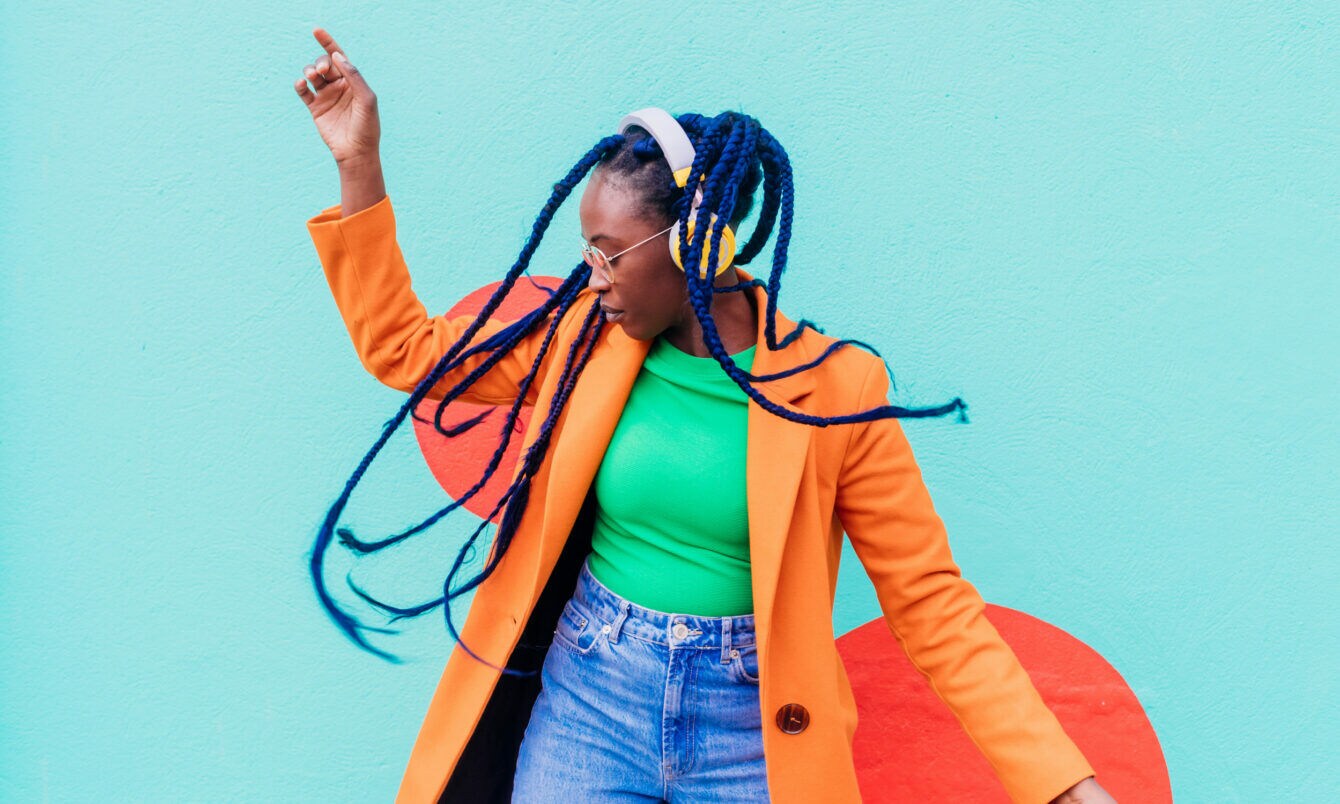Eine junge Frau mit blauen Rastazöpfen in einem grünen Shirt und orangefarbenen Mantel tanzt mit gelben Kopfhörern vor einer türkisfarbenen Fassade.