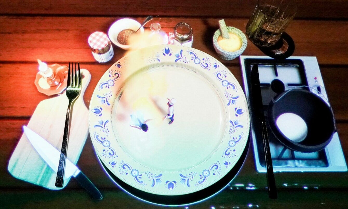 Ein gedeckter Tisch mit einem großen weißen Teller, auf dem zwei kleine Figuren stehen. Daneben befinden sich ein Messer, eine Gabel, ein Glas, Gewürzgläser und eine Pfanne mit einem Ei.