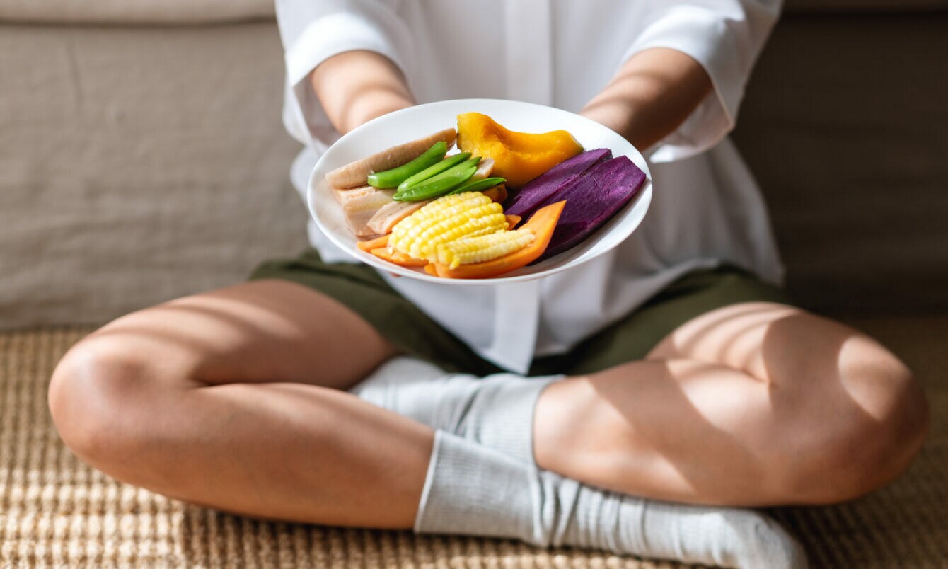 Person sitzt im Schneidersitz und hält einen Teller mit buntem, geschnittenem Gemüse, darunter Mais, Süßkartoffel und grüne Bohnen.