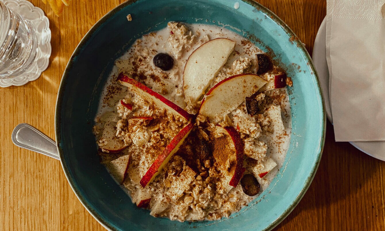 Blaue Schüssel mit Porridge, garniert mit Apfelscheiben, Blaubeeren und Zimt, auf Holztisch mit Glas Wasser und Serviette.