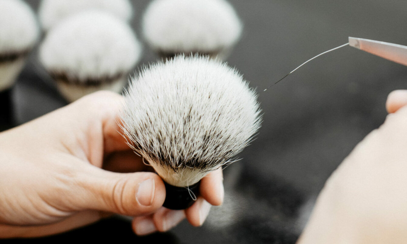 Hand hält einen weißen Rasierpinsel mit dunklen Borsten, eine zweite Hand führt eine Pinzette an die Borstenspitze.