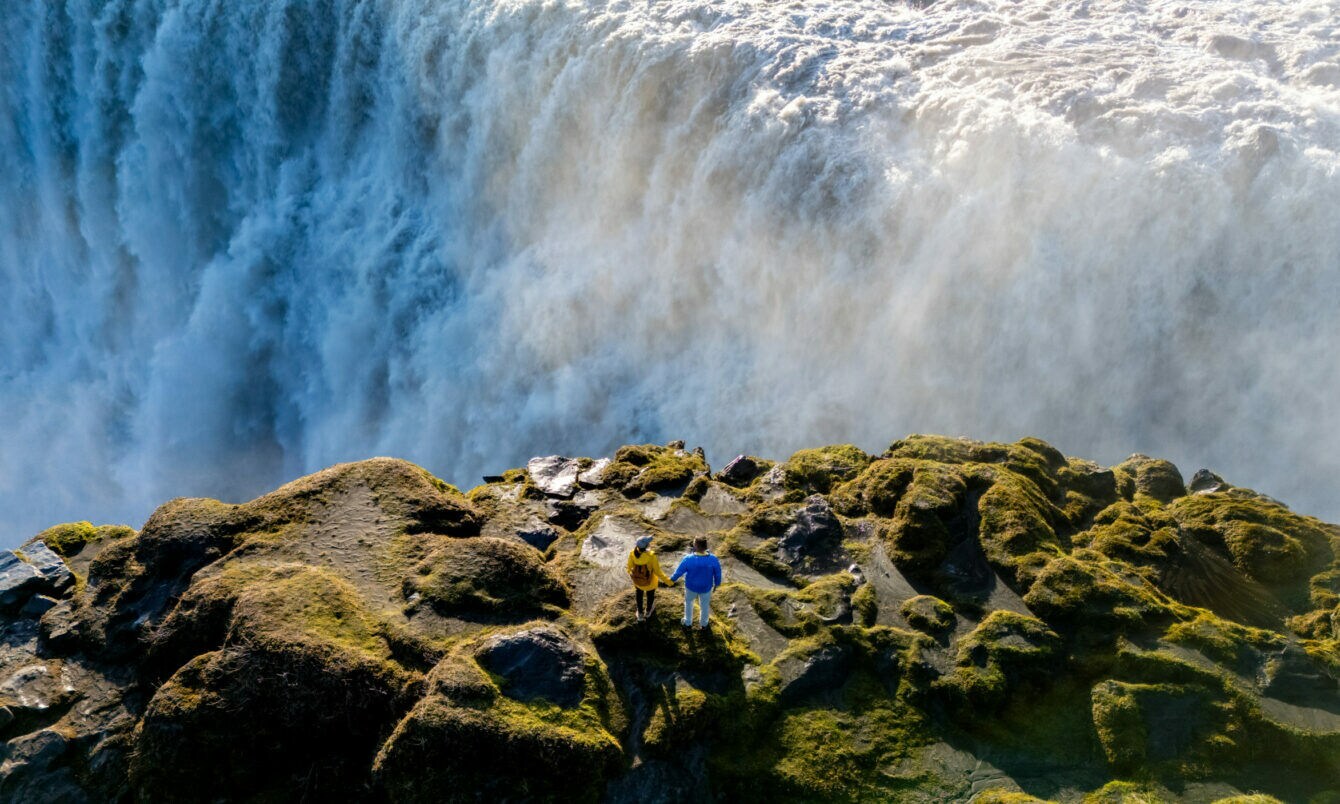 Zwei Personen stehen auf moosbedecktem Felsen vor einem großen Wasserfall mit starker Gischtentwicklung.