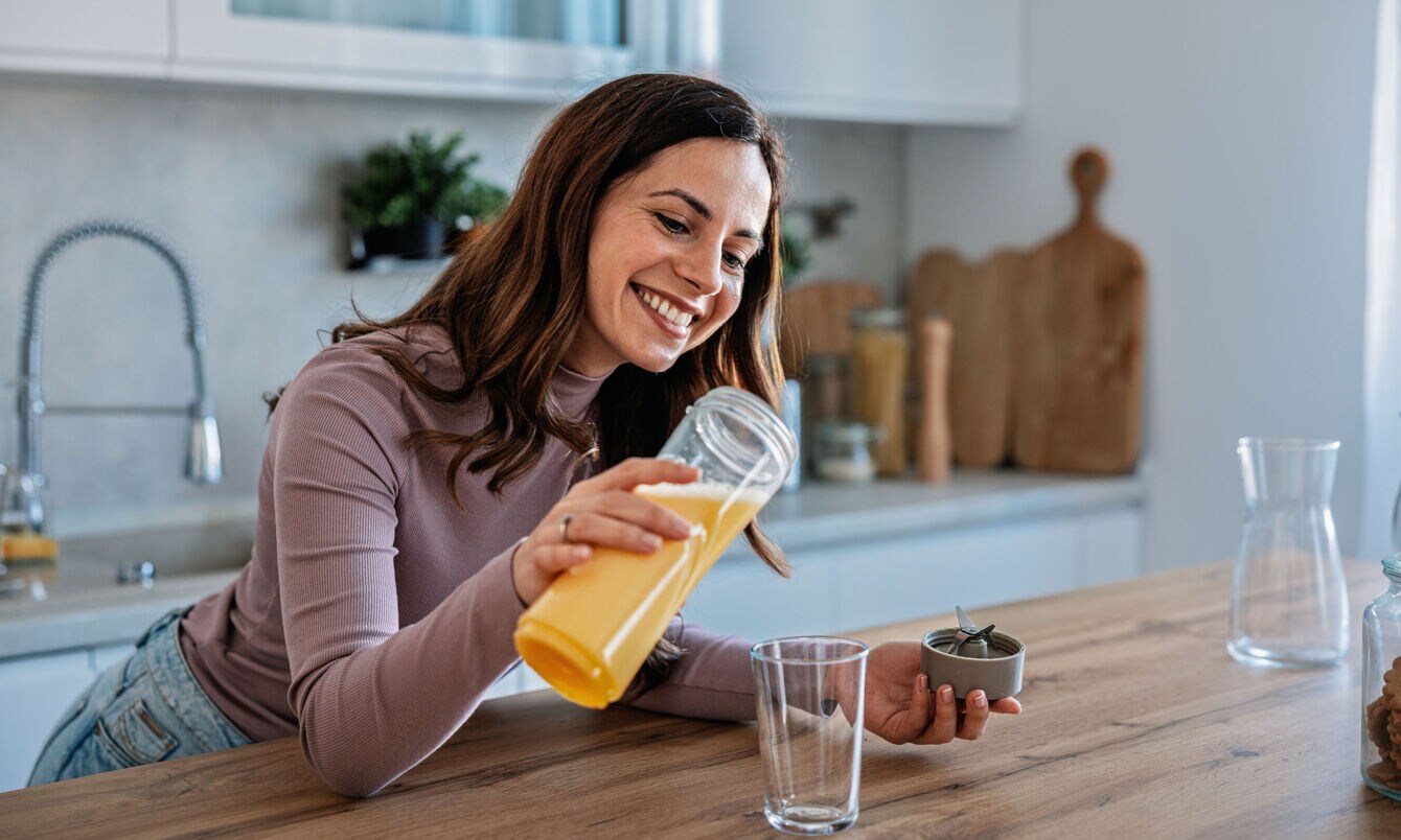 Eine lächelnde Frau gießt ein gelbes Getränk in eine Glas an einer Küchentheke.