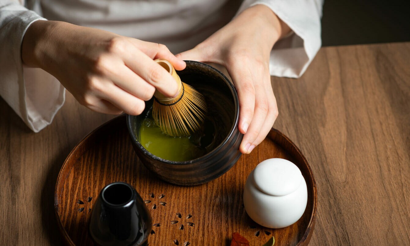 Nahaufnahme von zwei Händen bei der Zubereitung eines grünen Tees mit einem Bambusbesen in einer dunklen Keramikschale auf einem Holztisch.
