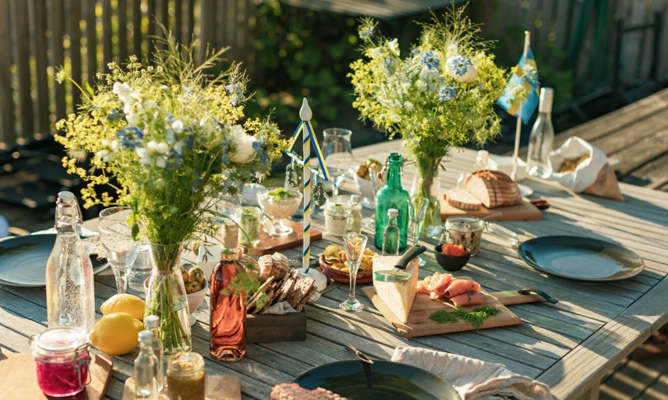 Gedeckter Holztisch für ein Mittsommer-Essen im Freien, mit Brot, gebeiztem Lachs, Käse, Eingelegtem, Getränken und frischen Blumensträußen.