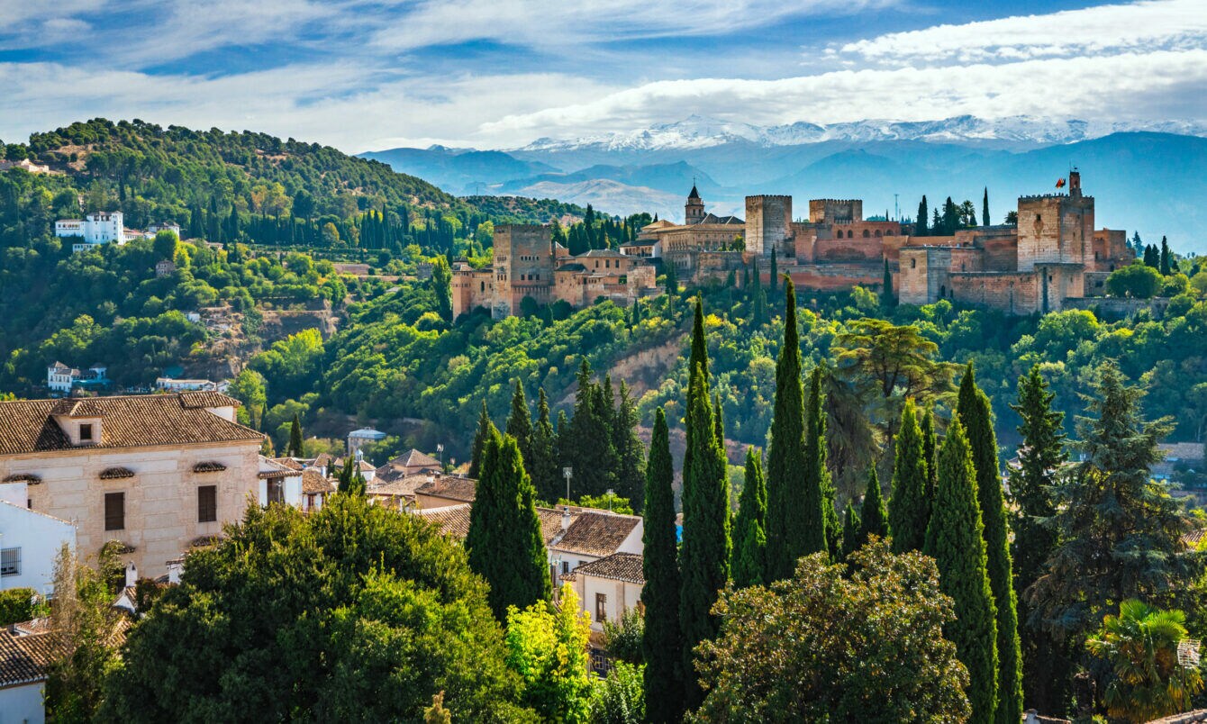 Blick auf einzelne Häuser und die von Wald umgebene Alhambra, im Hintergrund Berge.