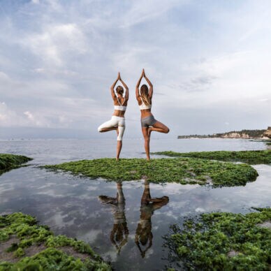 Zwei Frauen stehen am Wasser und machen zwei Yogaübungen