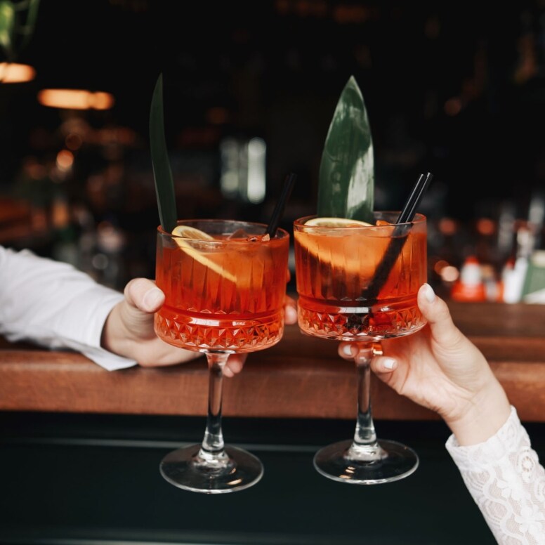 Zwei Spritz-Getränke mit Orangenscheiben in den Händen zweier Personen beim Anstoßen an einem Bartresen.