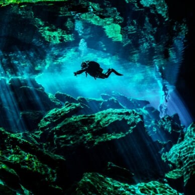 Eine Person in Taucherausrüstung taucht in einer von Wasser gefüllten Höhle. Lichtsäulen ergeben sich durch den starken Lichteinfall von oben im Wasser.