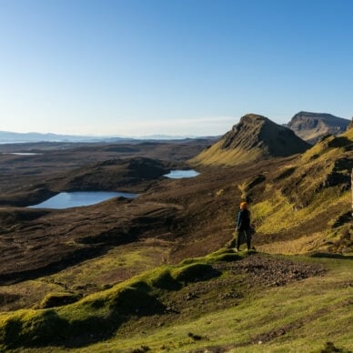 Eine Person steht in einer Landschaft aus Wiesen, Hügeln und Seen in Schottland. 