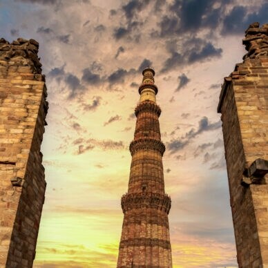 Das Qutub Minar, ein hoher roter Sandsteinturm, umgeben von Ruinenmauern bei Sonnenuntergang mit bewölktem Himmel