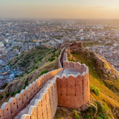 Luftaufnahme der historischen Stadtmauer von Jaipur, die sich über grüne Hügel erstreckt, mit der Stadt im Hintergrund bei Sonnenuntergang