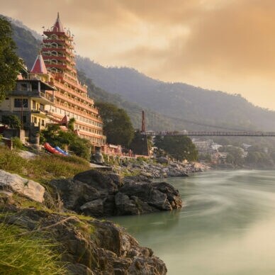 Fluss mit hängender Fußgängerbrücke und mehrstöckigem Tempelgebäude am Ufer bei Sonnenuntergang in bergiger Landschaft