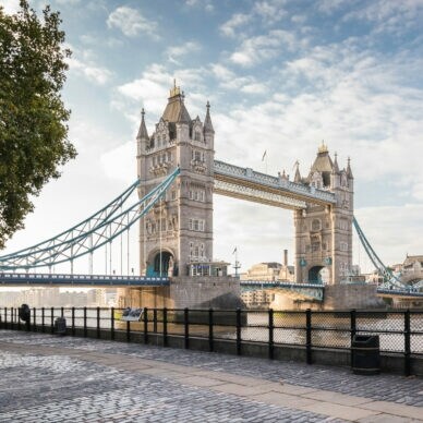 Tower Bridge über der Themse mit zwei markanten Türmen und blau-weißen Hängebrückenabschnitten, Blick vom Ufer mit Bäumen und Laterne.