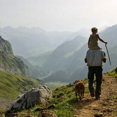 Ein Erwachsener trägt ein Kind auf den Schultern, während sie einen Wanderweg entlanggehen. Ein Hund läuft neben ihnen. Die Landschaft zeigt Berge und Täler im Hintergrund.