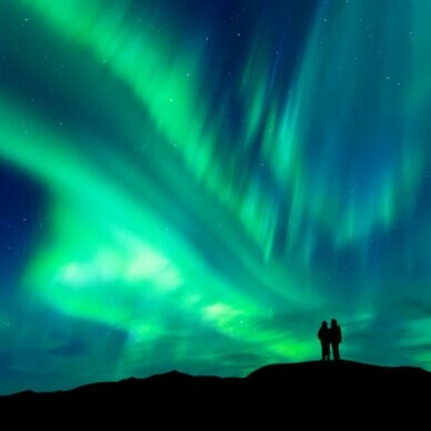 Zwei Personen stehen auf einem Hügel und beobachten das grün leuchtende Nordlicht am Nachthimmel in Norwegen.