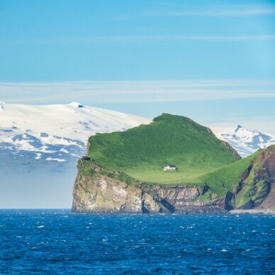 Blick auf die grüne isländische Insel Ellidaey mit einer einsamen Jagdhütte darauf und dem, unter einem Gletscher liegenden Vulkan Eyjafjallajökull im Hintergrund.