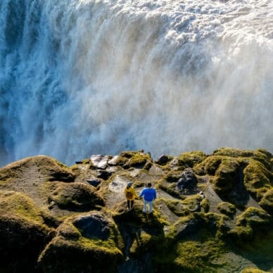 Zwei Personen stehen auf moosbedecktem Felsen vor einem großen Wasserfall mit starker Gischtentwicklung.