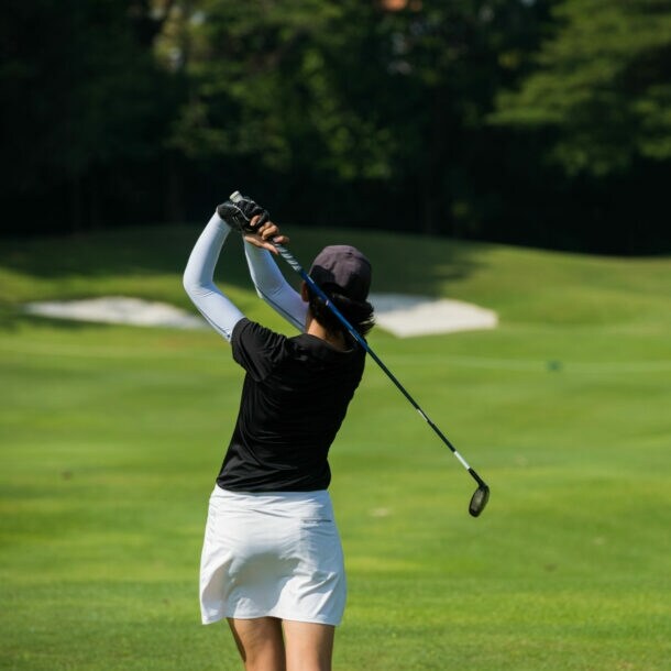 Golferin von hinten holt zum Schlag aus und sielt Richtung Topfbunker vor Bäumen.