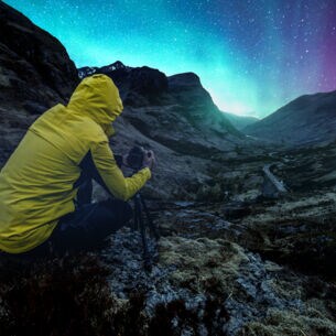 Eine Person in wasser- und windabweisender Kleidung fotografiert einen lila-blau leuchteten Sternenhimmel