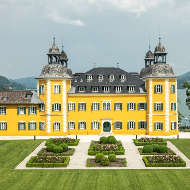Ein gelbes Schlosshotel vor dem Panorama des Wörthersees