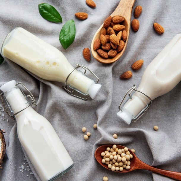 Auf einem grauen Tuch liegen drei Bügelflaschen mit weißlicher Flüssigkeit, Kokosraspel, Kichererbsen und Mandeln