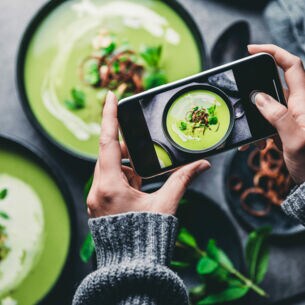 Zwei Hände halten Smartphone, das eine grüne Suppe in Schüssel fotografiert
