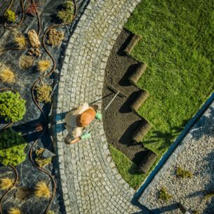 Luftaufnahme einer Person, die auf einem gepflasterten Weg stehend eine Fläche mit Rollrasen in einem Garten bearbeitet