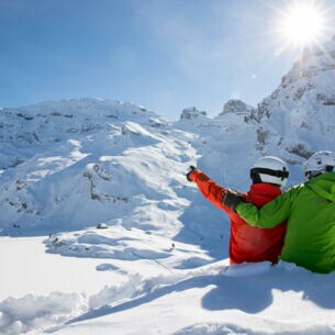 Zwei Skifahrer:innen von hinten im Schnee, sie blicken auf die Berggipfel