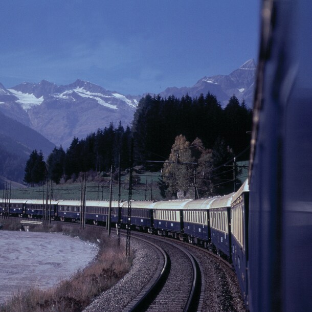 Ein blauer Zug fährt in einer Kurve in der Dämmerung durch die Landschaft, im Hintergrund Wald und Bergpanorama