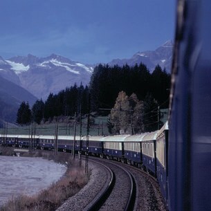Ein blauer Zug fährt in einer Kurve in der Dämmerung durch die Landschaft, im Hintergrund Wald und Bergpanorama